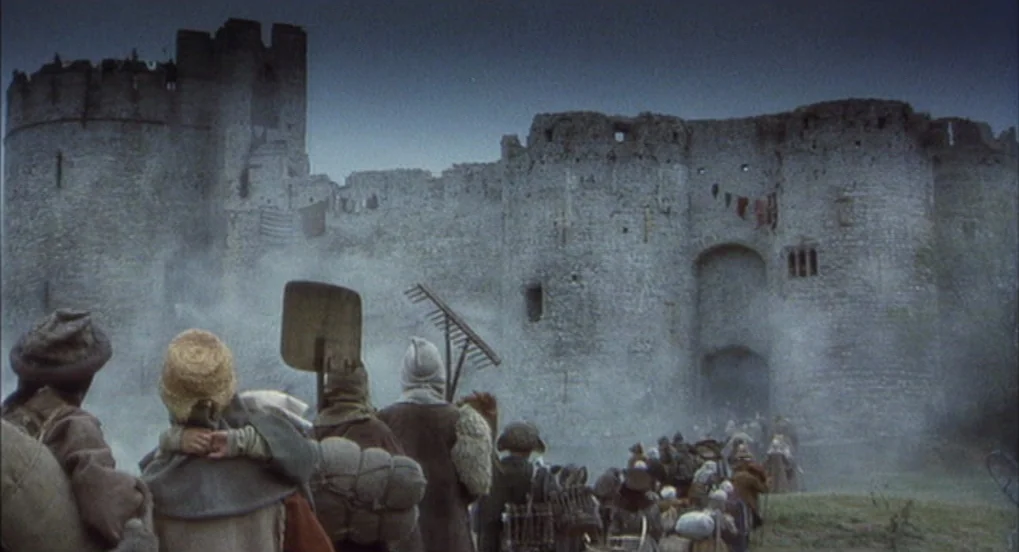 Jabberwocky (1977) movie still: wide — A massive grey stone castle with circular towers stands amidst a hazy, overcast day. A large crowd of people…