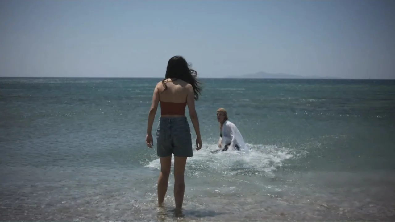 Hot Milk (2025) movie still: fullbody — A sunny coastal shoreline with calm water meeting the sandy ground under a clear sky. A woman stands in the…
