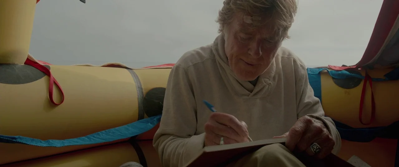 All Is Lost (2013) movie still: medium — An elderly man sits inside a yellow rubber inflatable raft on an overcast day. He is writing in a small…