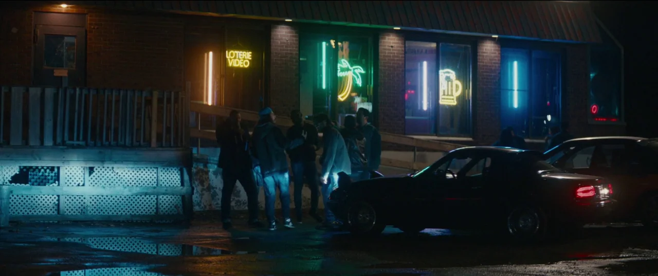 Humanist Vampire Seeking Consenting Suicidal Person (2023) movie still: wide — A brick storefront at night features glowing neon signage including a…