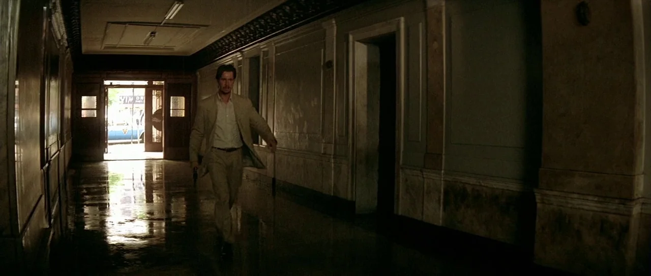 Léon: The Professional movie still: fullbody — A long, shadowed office building hallway has polished floors reflecting light from a doorway. A man…