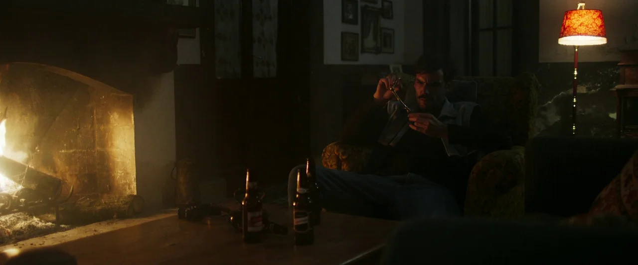 When Evil Lurks (2023) movie still: wide — A dimly lit room features a glowing fireplace and an ornate floor lamp nearby. A man sits in a patterned…