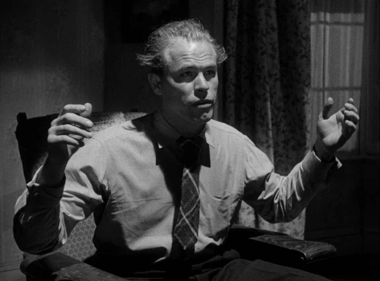 The Killers (1946) movie still: medium — A dimly lit room with an armchair placed near a window with curtains. A man sits in the chair with his…