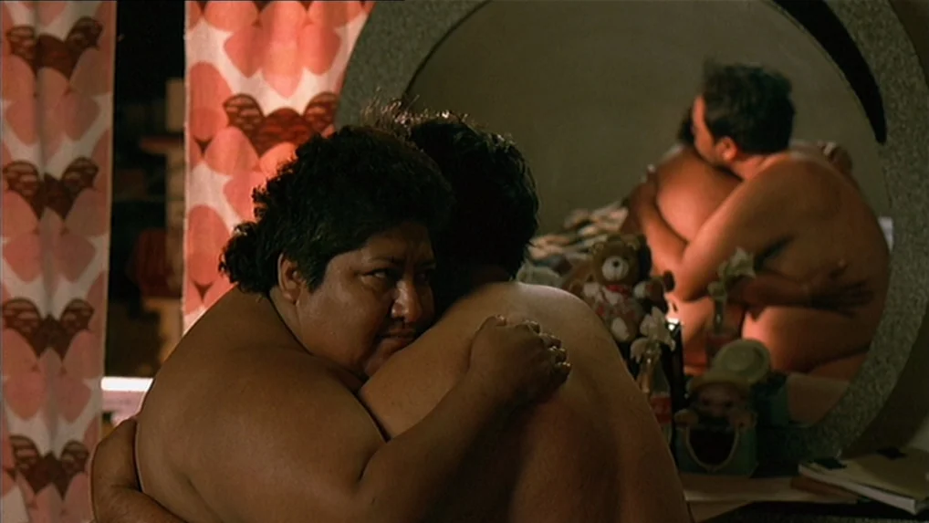 Battle In Heaven (2005) movie still: medium — A dimly lit room features a large circular mirror reflecting a couple hugging. The woman faces the…