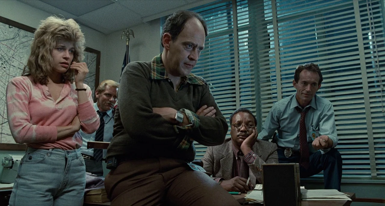 The Terminator (1984) movie still: medium — A cramped, dimly lit workspace filled with blinds and administrative files. Several people stand in the…
