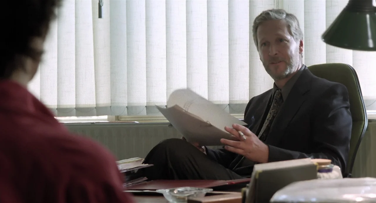 Thesis (1996) movie still: medium — An office features vertical window blinds and a desk cluttered with documents and papers. A man in a suit sits…