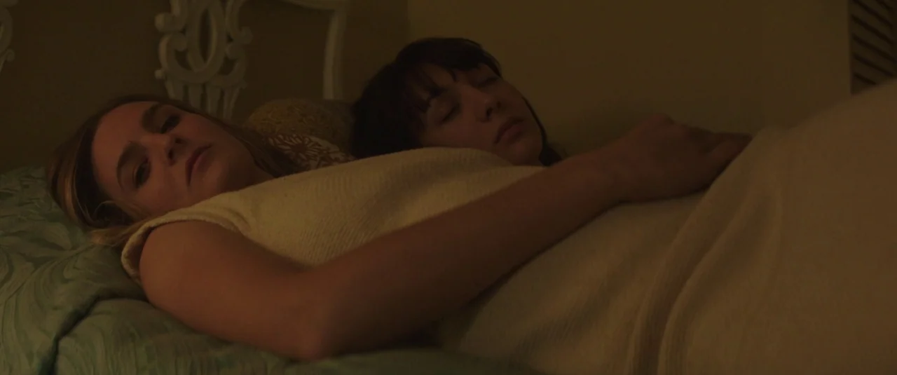 Southbound (2015) movie still: medium — Two women lie together on a bed covered in green-patterned bedding and white linens. One woman is wide awake…