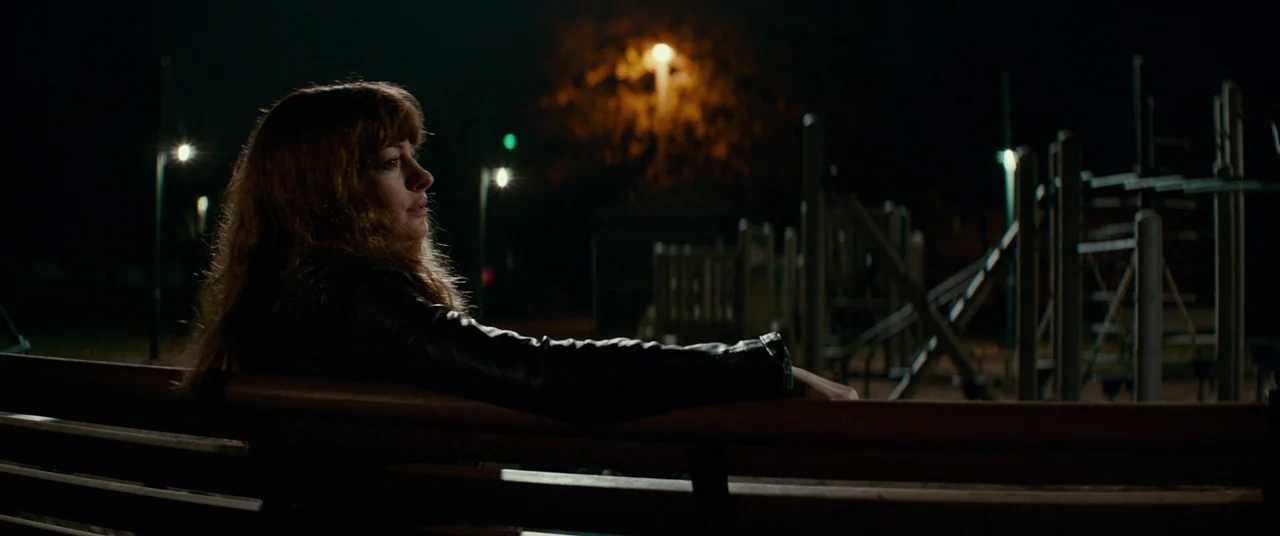 Colossal (2016) movie still: medium — A dimly lit park at night features a wooden bench in the foreground. A woman with long hair sits on the bench…