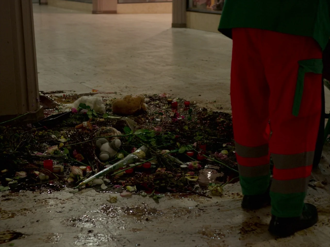 Violet (2014) movie still: medium — A tiled indoor floor features a pile of flowers, stuffed toys, and spilled liquid. A person in a work uniform…