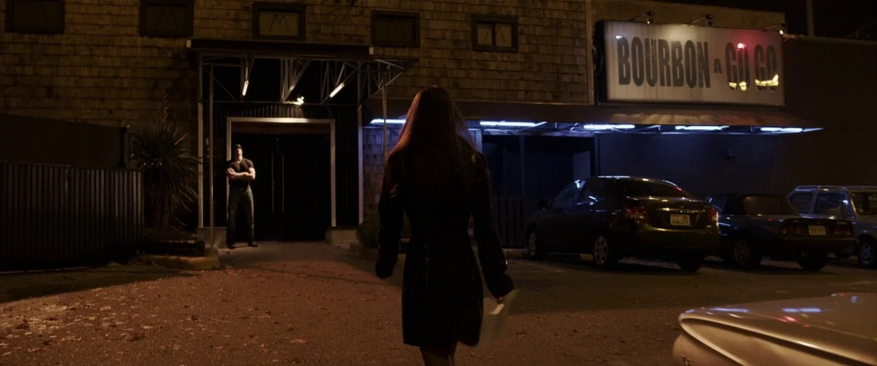 American Mary (2012) movie still: wide — A dimly lit parking lot at night sits in front of a shingled building structure. A woman approaches the…