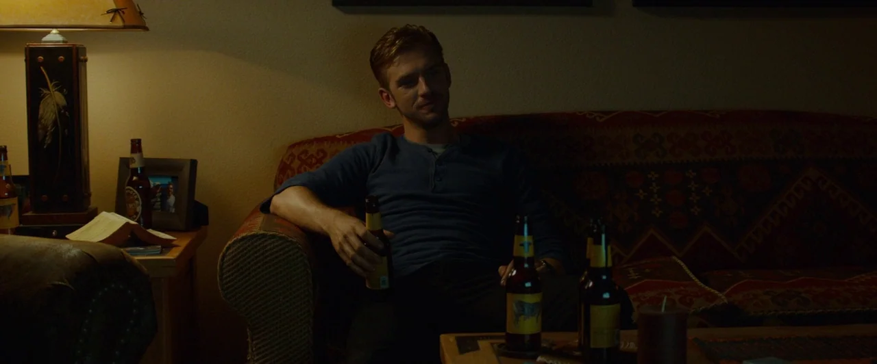 The Guest (2014) movie still: medium — A dimly lit room features a patterned sofa against a dark wall. A man sits with a beer bottle in hand near a…