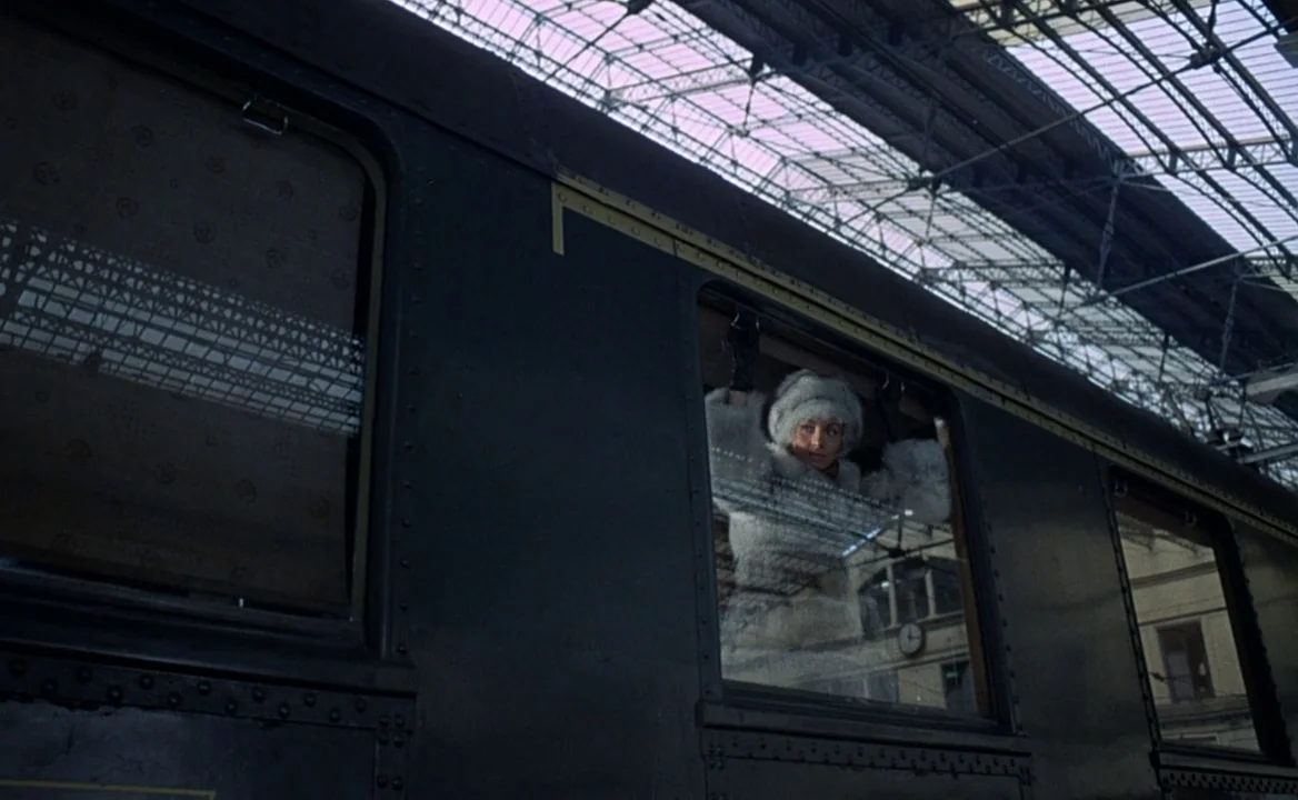 F for Fake (1973) movie still: wide — A dark green train carriage sits under the arched steel frame of a station. A woman in a white fur hat looks…