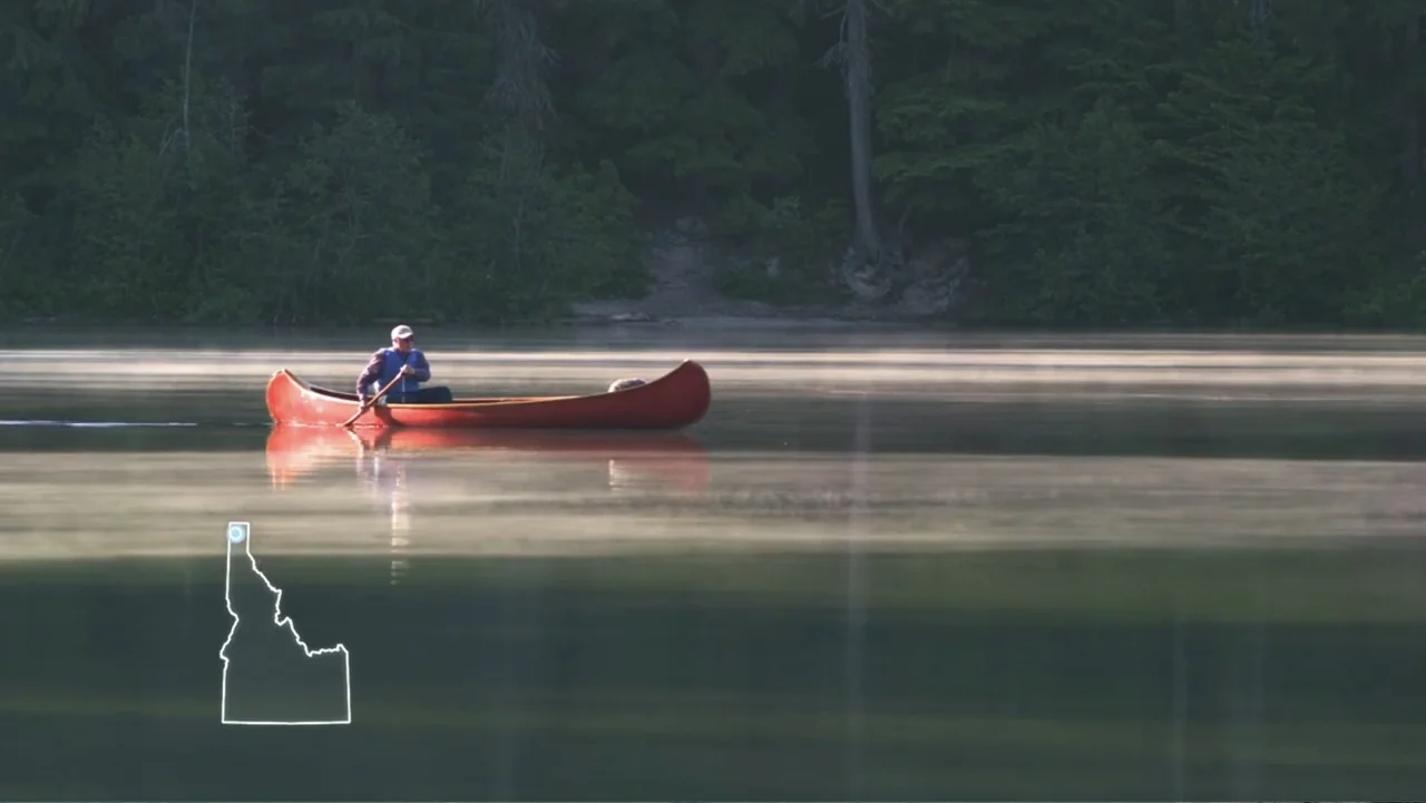 Idaho: The Movie (2012) movie still: wide — A calm lake is covered in a layer of low-hanging morning mist. A man paddles a red canoe across the…