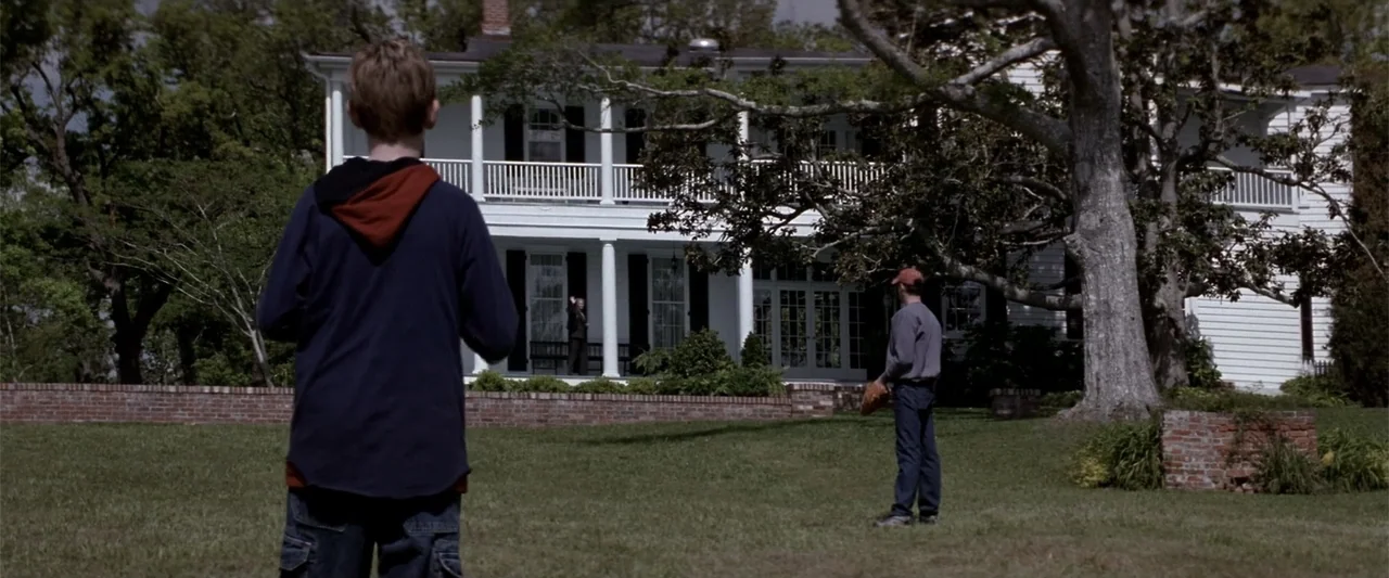 Angst (2017) movie still: wide — A large two-story white colonial house features a wrap-around porch and black shutters. A boy and a man stand on…