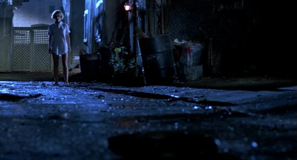 A Nightmare On Elm Street (1984) movie still: fullbody — A dark city alleyway at night features metal trash bins and a chain-link fence. A young…