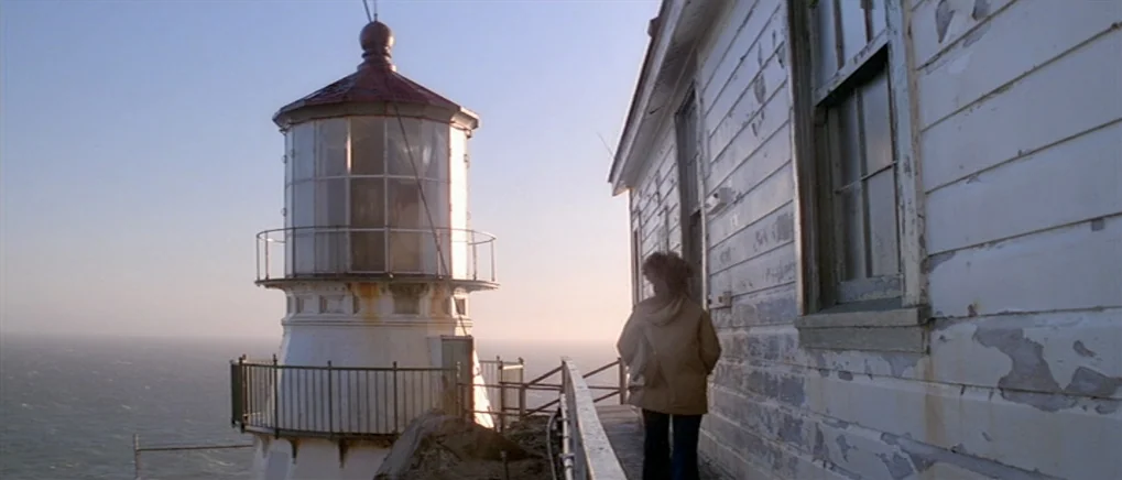 The Fog (1980) movie still: wide — A white lighthouse with peeling paint sits beside a wooden structure overlooking the ocean. A woman walks along…