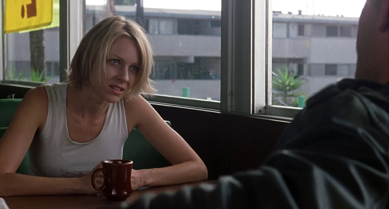 Mulholland Drive (2001) movie still: medium — A woman sits at a table inside a diner booth near large windows. She gazes at a man whose back is…