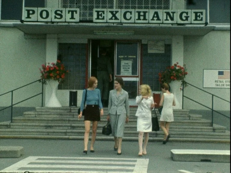 Der Bomberpilot (1970) movie still: wide — A Post Exchange building facade features concrete steps, large signs, and potted red flowers. Four women…
