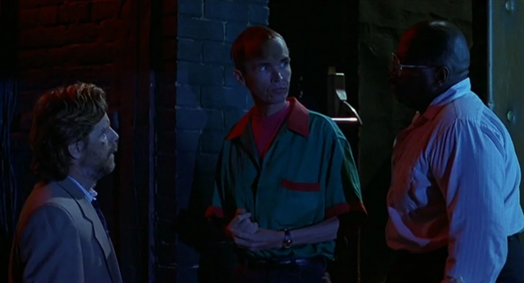 Exotica (1994) movie still: medium — An exterior nighttime alleyway features a dark brick wall backdrop. Three men stand in a cluster while…