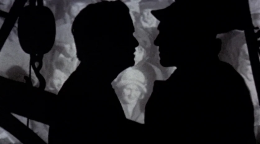The Saddest Music in the World (2003) movie still: medium — A dark space features two figures in silhouette against a grainy projection of people. A…