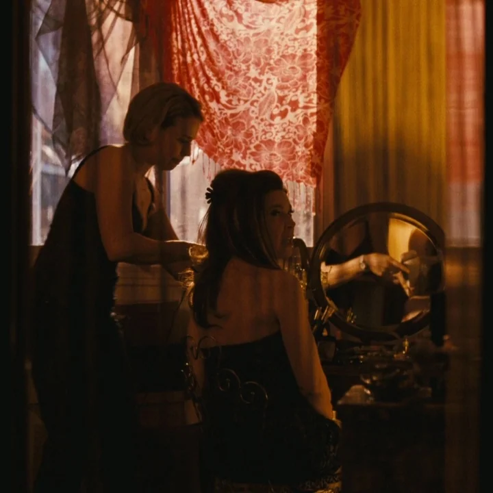 Mommy (2014) movie still: medium — A dimly lit room with patterned red fabric draped near a window. One woman stands behind a seated woman who faces…