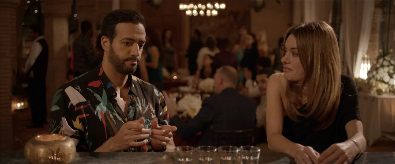 An Endless Wedding (2025) movie still: medium — A dimly lit restaurant bar features a counter with glassware and blurred background guests. A man…