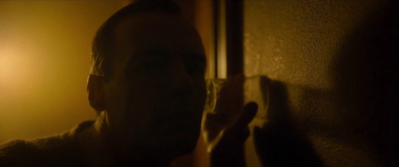 Earwig (2022) movie still: closeup — An interior space features a textured wall under warm low-key light. A man holds a glass against the wall to…