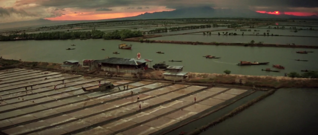 Apocalypse Now (1979) movie still: establishing — Vast, partitioned salt evaporation ponds stretch toward a wide river under a dark sky. A military…