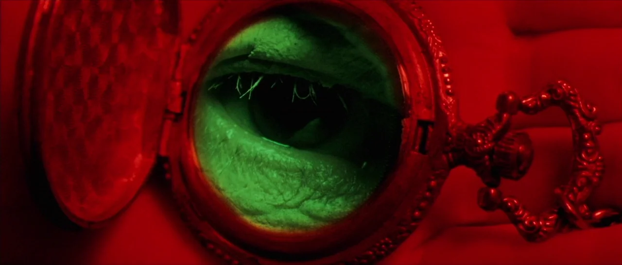Amer (2009) movie still: closeup — A close-up view of an antique pocket watch held within a red-lit environment. A human eye is clearly visible in…