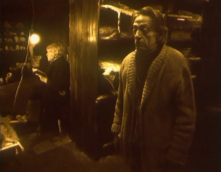 Dead Man’s Letters (1986) movie still: medium — A dim, cluttered room contains a man standing near a tall cabinet in the foreground. A second person…