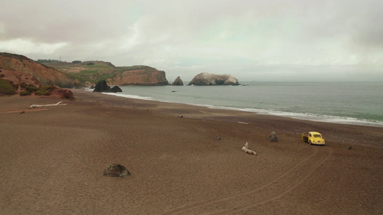 Bumblebee (2018) movie still: wide — A wide, deserted beach lies beneath a pale, overcast sky next to dark cliffs. A single yellow compact car is…