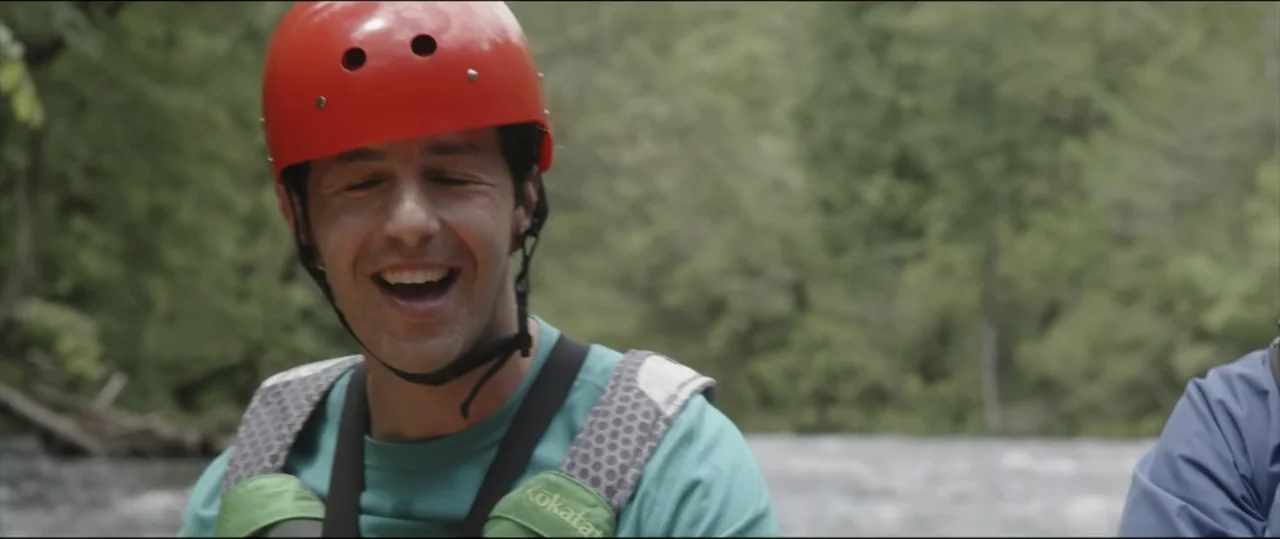 Summer Camp (2024) movie still: medium — A man in a river setting wears a bright red helmet and life vest. He smiles broadly while looking toward…