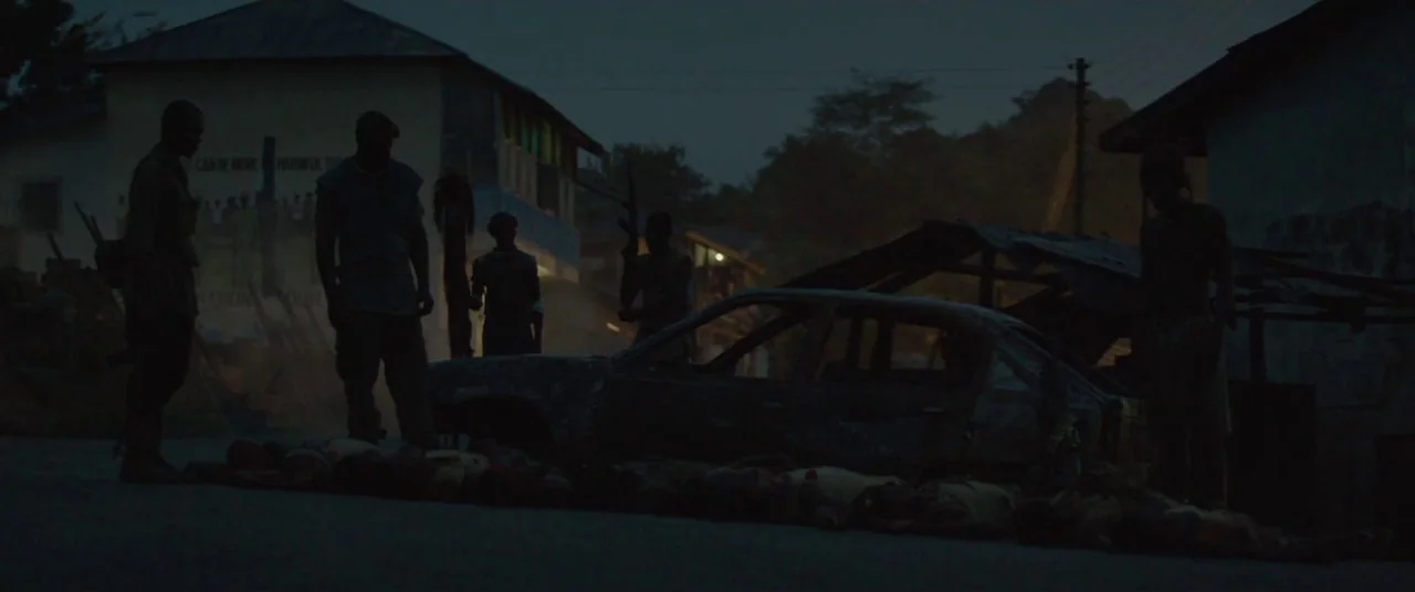 Beasts of No Nation (2015) movie still: wide — A dark, dilapidated village street features small buildings and a burnt-out vehicle carcass. Several…