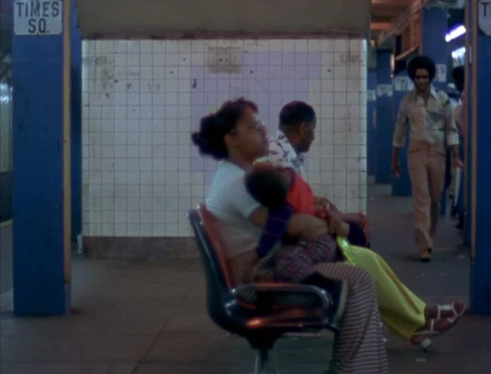 News From Home (1977) movie still: fullbody — A Times Square subway platform features white tiled walls and blue-painted steel support pillars. A…