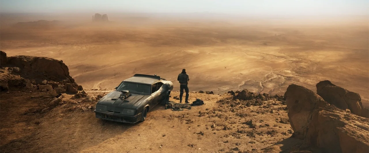 Furiosa: A Mad Max Saga (2024) movie still: wide — A vast, arid desert landscape features distant rock formations under a dusty hazy sky. A man…