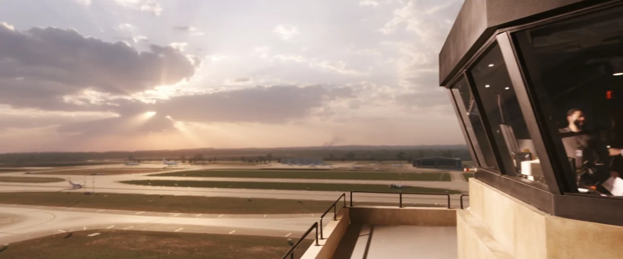 On a Wing and a Prayer (2023) movie still: wide — An airport control tower overlooks wide flat runways under a dramatic golden hour sky. Clouds…