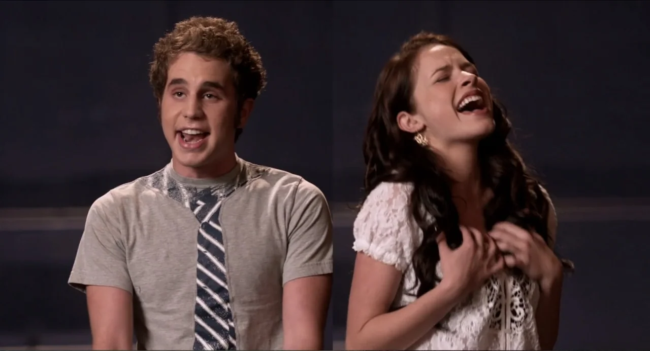 Pitch Perfect (2012) movie still: medium — Two performers stand side-by-side against a plain dark wall during a vocal performance. The man sings…