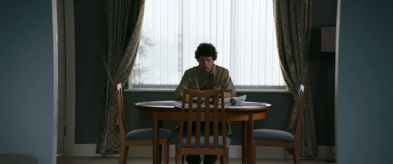 Vivarium (2019) movie still: medium — A man sits at a round wooden dining table centered before a window with curtains. He eats his meal alone in a…