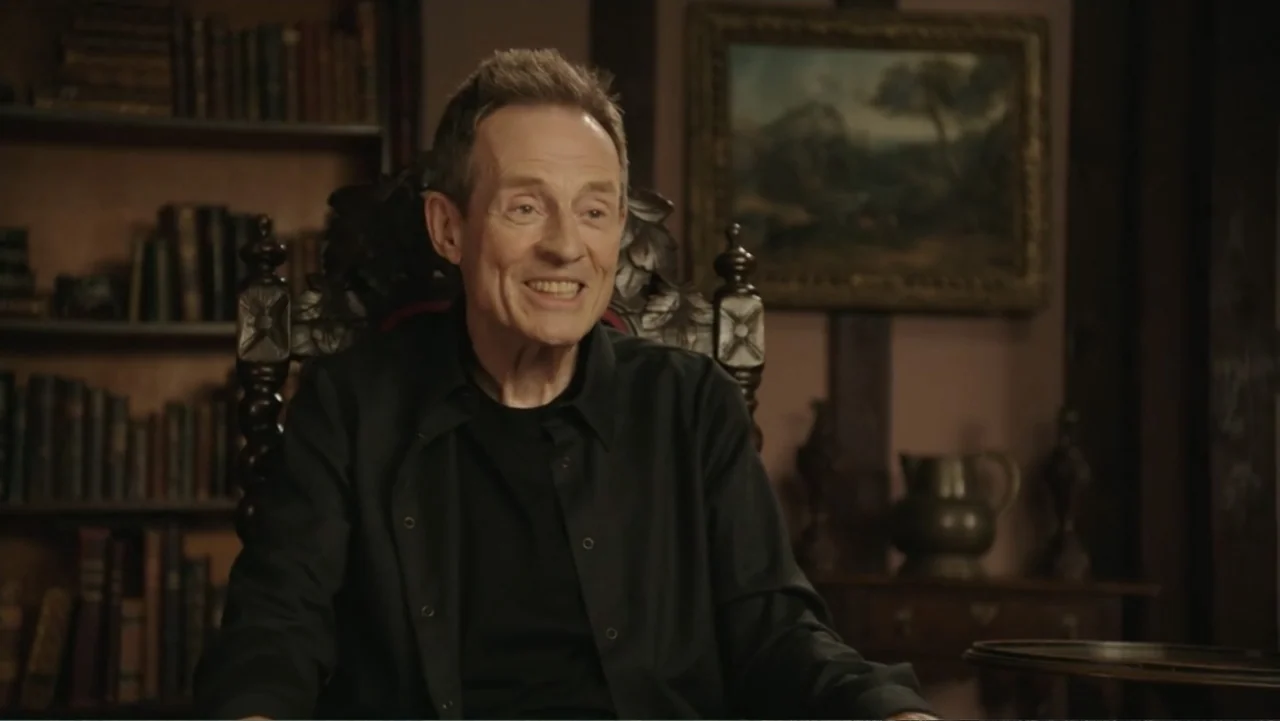 Becoming Led Zeppelin (2025) movie still: medium — A dimly lit room with rows of leather-bound books and a framed landscape painting. An older man…