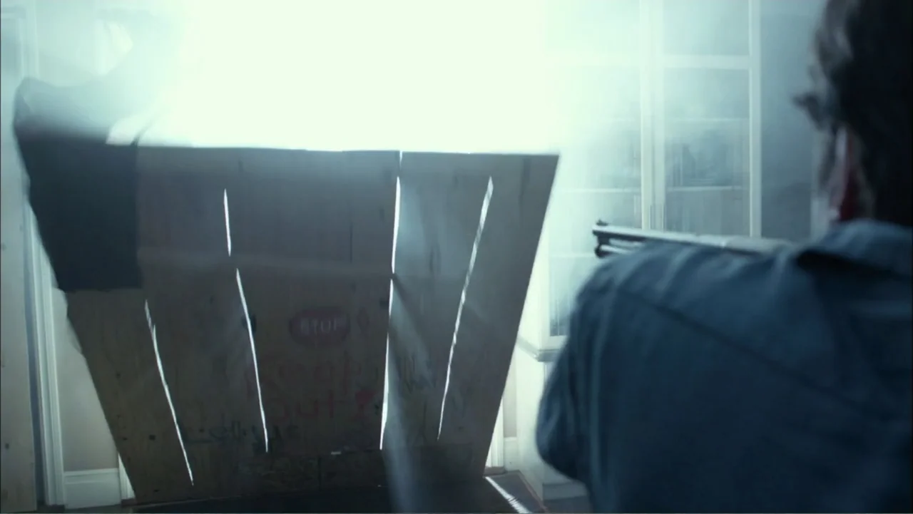 Dark Skies (2013) movie still: medium — A bright doorway is partially blocked by a piece of plywood with graffiti markings. A man holding a shotgun…