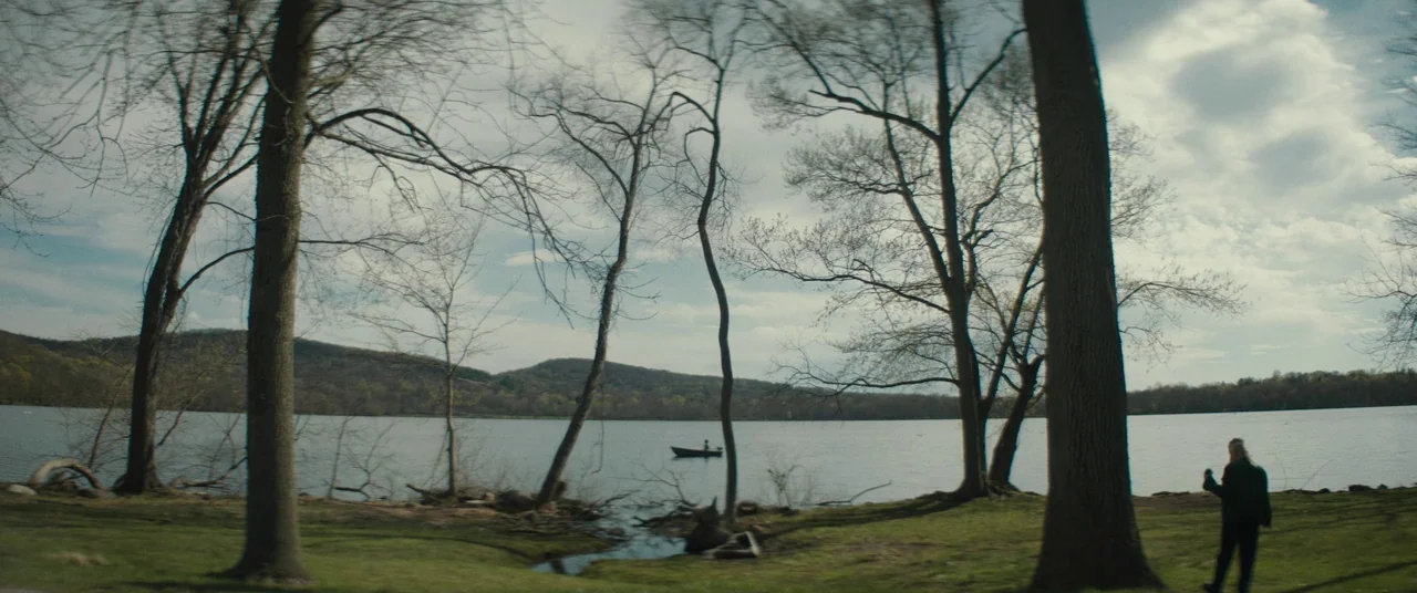 I Think We’re Alone Now (2018) movie still: wide — A wide view of a calm lake surrounded by leafless trees under a cloudy sky. A person stands near…