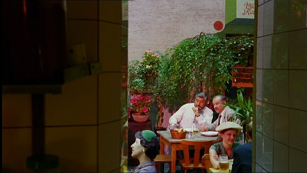 Lola (1981) movie still: wide — An outdoor restaurant patio features several tables surrounded by lush potted greenery and trees. Several…