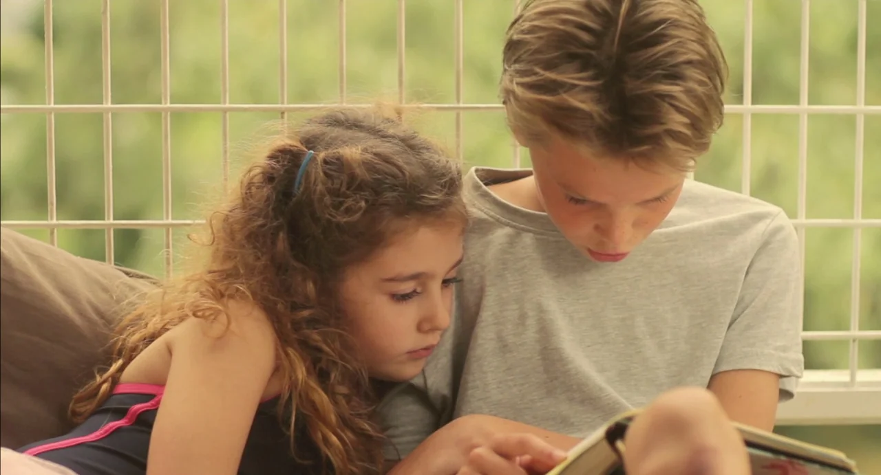 Tomboy (2011) movie still: medium — Two children lean against a white lattice screen on a bright, outdoor-facing balcony. The girl rests her head…