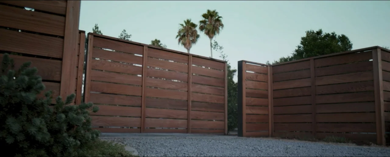 Hostages (2017) movie still: establishing — A wooden horizontal slat fence stands before a gravel driveway with tall palm trees. No occupants are…