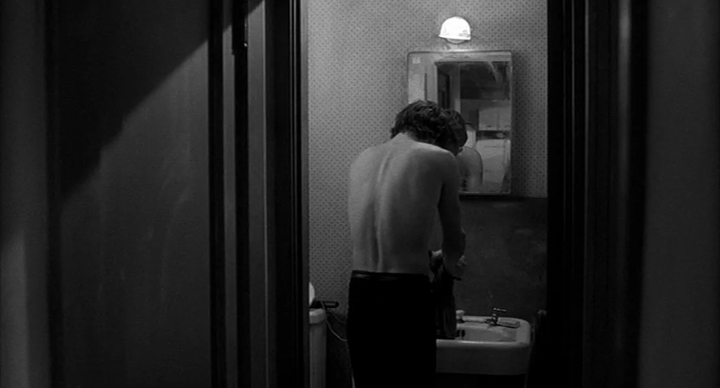 Rumble Fish (1983) movie still: medium — A small bathroom with patterned wallpaper features a mirror and a porcelain sink. A man stands shirtless…