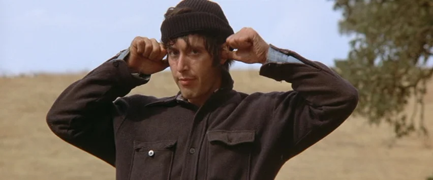 Scarecrow (1973) movie still: medium — A dry, grassy field serves as the backdrop under a clear sky. A man wears a beanie and a dark brown jacket…