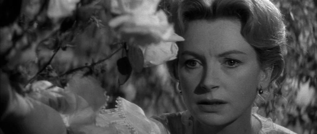 The Innocents (1961) movie still: closeup — A woman stands beside a dense bush filled with large white blossoms. She reaches a hand toward the…