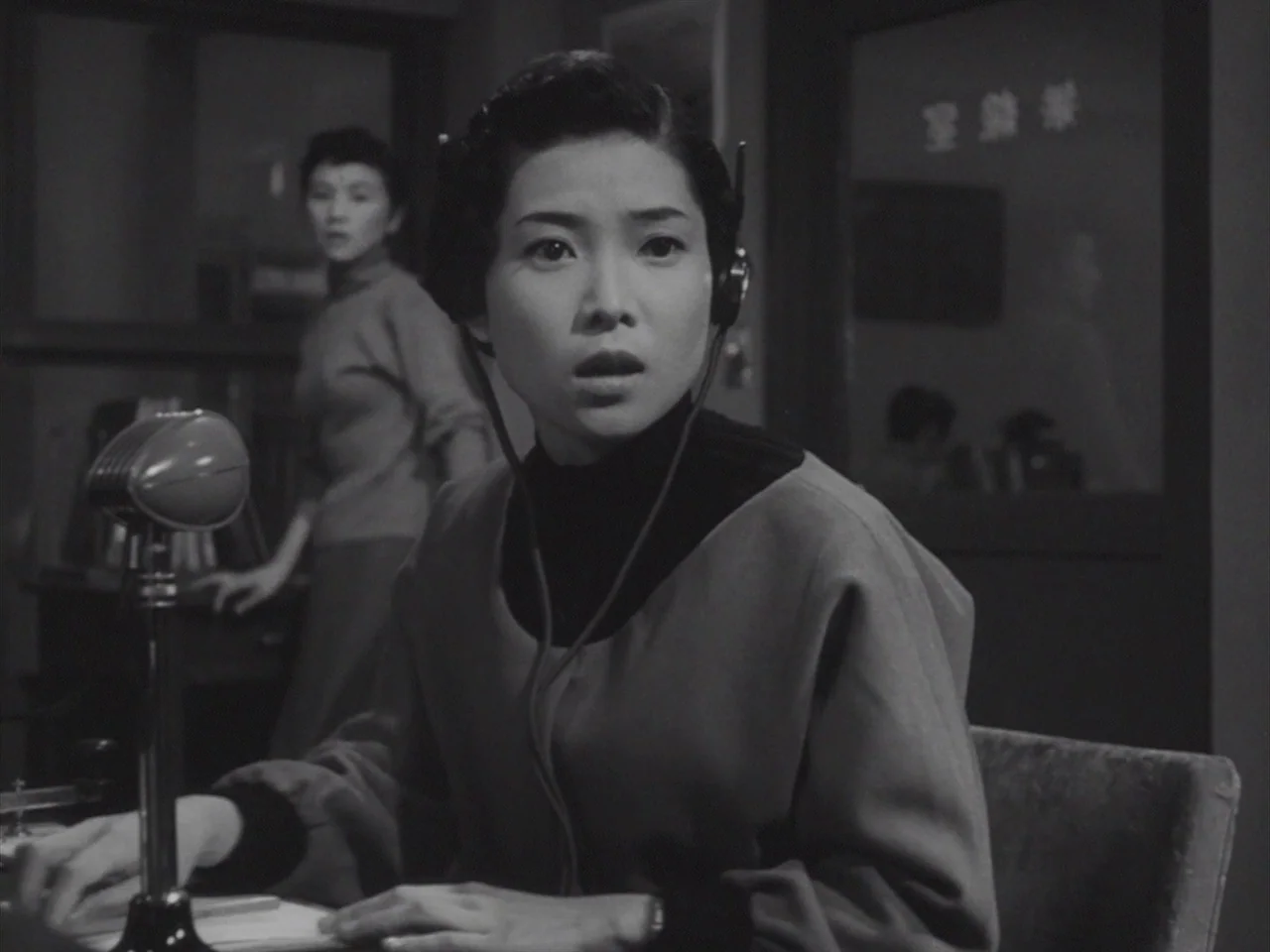 Godzilla Raids Again (1955) movie still: medium — A radio studio with a metal microphone on a desk and glass partitions visible. A woman in a…