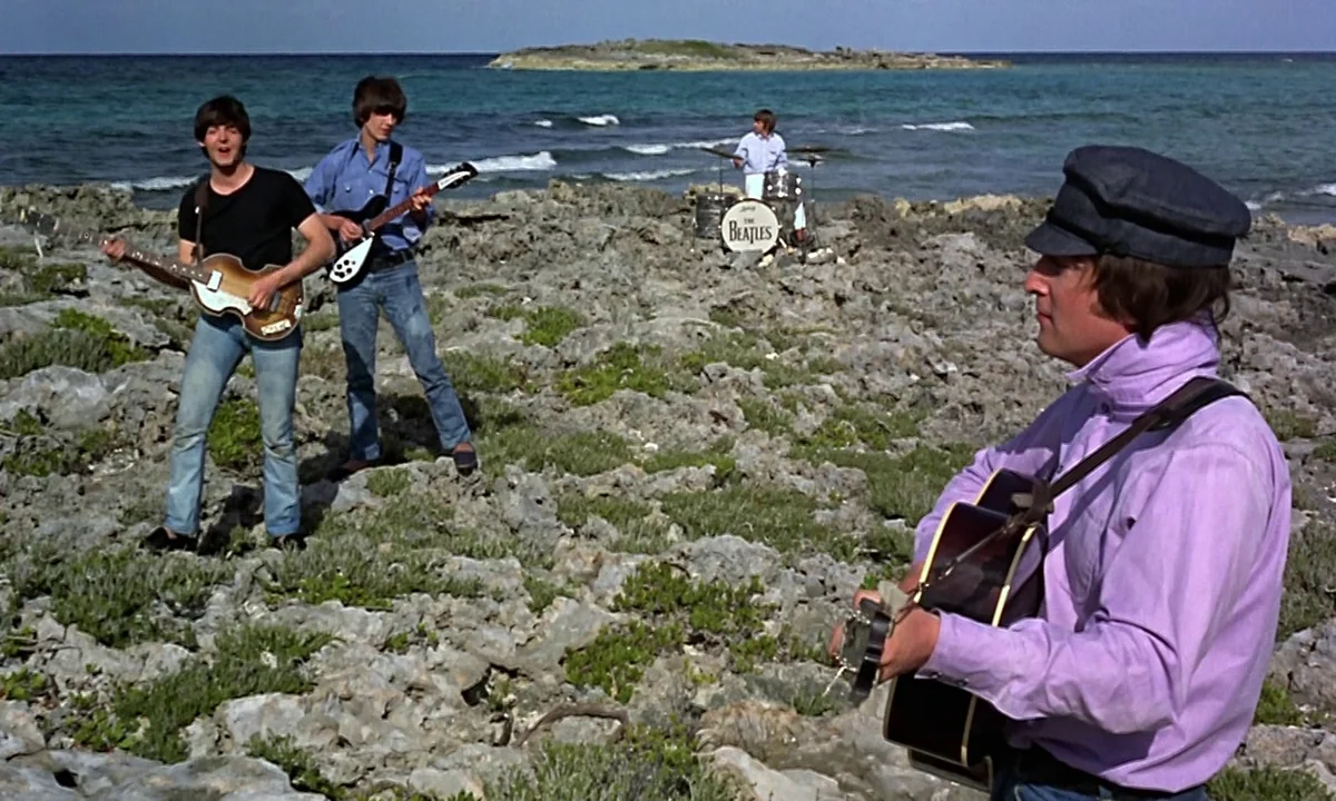 Help! (1965) movie still: wide — A rocky shoreline sits beside a calm sea under a clear blue sky. Four men in casual clothing perform on guitars and…