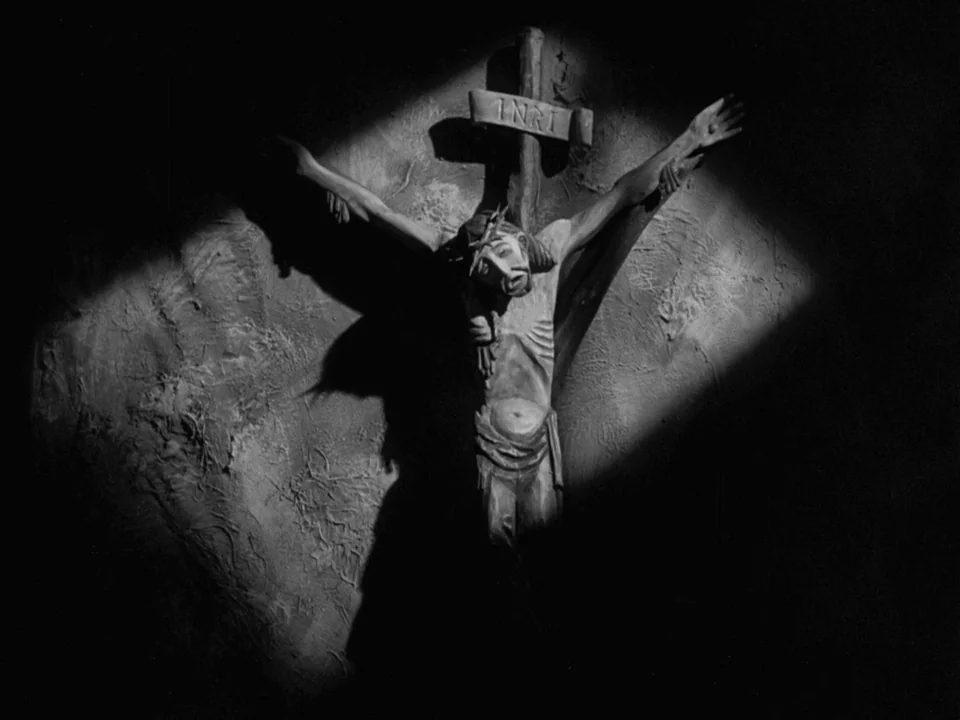 The Virgin Spring (1960) movie still: closeup — A wooden crucifix is mounted on a textured, rough-plastered wall in deep darkness. A spotlight…